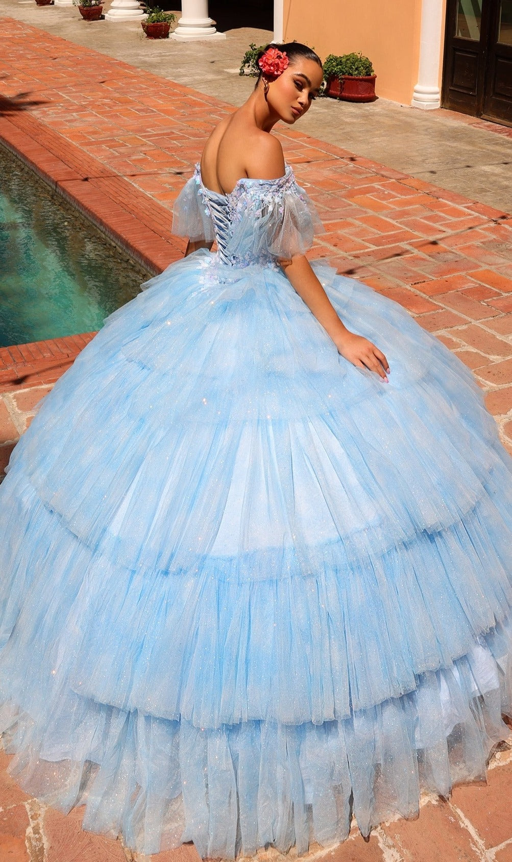A woman in the Quinceanera Dress 54312 by Amarra, a light blue off-the-shoulder ball gown with layered tulle, stands beside a pool on a brick patio, looking over her shoulder.
